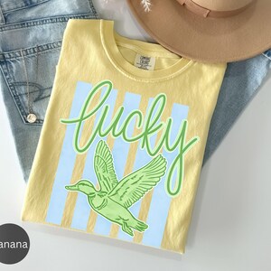May include: A yellow t-shirt with the word "lucky" in green script and a green duck graphic. The shirt has light blue vertical stripes and is folded on top of a pair of blue jeans and a tan hat.