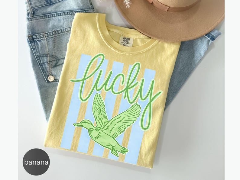 May include: A yellow t-shirt with the word "lucky" in green script and a green duck graphic. The shirt has light blue vertical stripes and is folded on top of a pair of blue jeans and a tan hat.