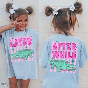 Puede incluir: Camiseta azul claro con un dise&ntilde;o de cocodrilo verde y rosa. La parte delantera de la camiseta dice "LATER" y la trasera "AFTER WHILE". Corte holgado, ideal para ni&ntilde;os.