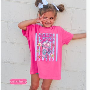 May include: A bright pink t-shirt with a graphic of a drink with flowers and the text "Dilly Dally". The shirt is worn by a young person with white bows in their hair.