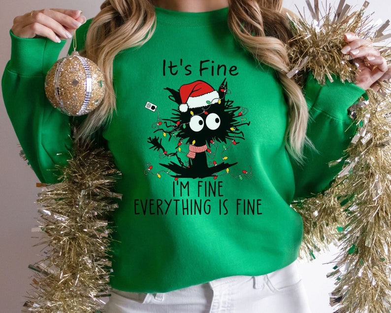 May include: A green Christmas sweater with a black cat wearing a Santa hat and tangled in Christmas lights. The text on the sweater reads 'It's Fine', 'I'm Fine', and 'Everything is Fine'.