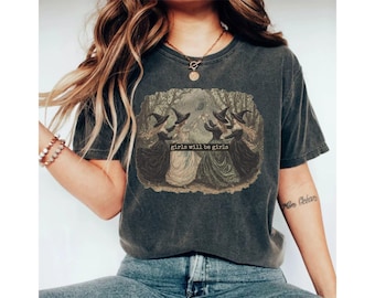 Könnte beinhalten: Dunkelgraues T-Shirt mit einer Illustration im Vintage-Stil von vier Hexen in schwarzen Hüten und Kleidern, mit dem Text "girls will be girls". Das Kunstwerk ist vor einem Waldhintergrund platziert.