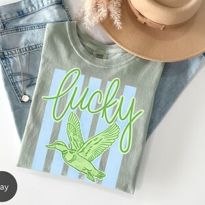 May include: A light green t-shirt with the word "lucky" in a script font and a green duck graphic. The shirt is laid on top of blue jeans and a tan hat.