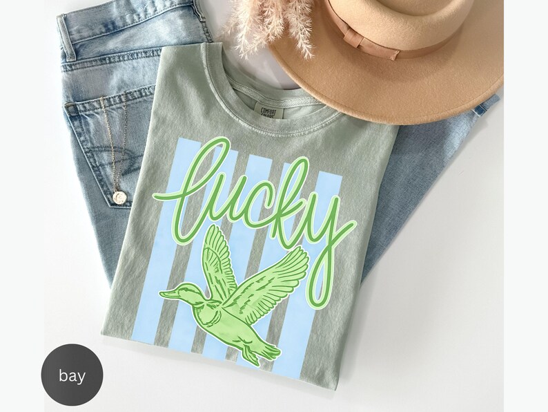 May include: A light green t-shirt with the word "lucky" in a script font and a green duck graphic. The shirt is laid on top of blue jeans and a tan hat.