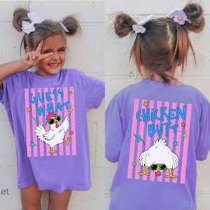 May include: A violet t-shirt with a graphic of a chicken wearing sunglasses and the text "GUESS WHAT" on the front. The back of the shirt features the text "CHICKEN BUTT" and a chicken bending over.