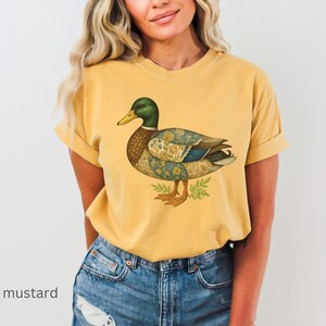 Puede incluir: Camiseta de color mostaza con una ilustraci&oacute;n detallada de un pato. El pato tiene una cabeza verde, un pico amarillo y un cuerpo estampado en tonos azules, marrones y dorados. La camiseta se combina con vaqueros azules.