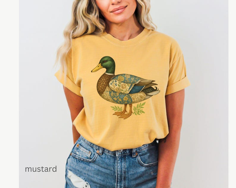 Puede incluir: Camiseta de color mostaza con una ilustraci&oacute;n detallada de un pato. El pato tiene una cabeza verde, un pico amarillo y un cuerpo estampado en tonos azules, marrones y dorados. La camiseta se combina con vaqueros azules.