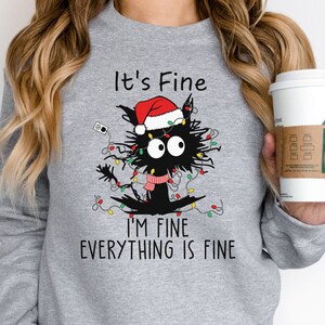 May include: A light gray sweatshirt featuring a black cat tangled in Christmas lights and wearing a Santa hat and scarf. The text "It's Fine, I'm Fine, Everything is Fine" is printed below the cat.