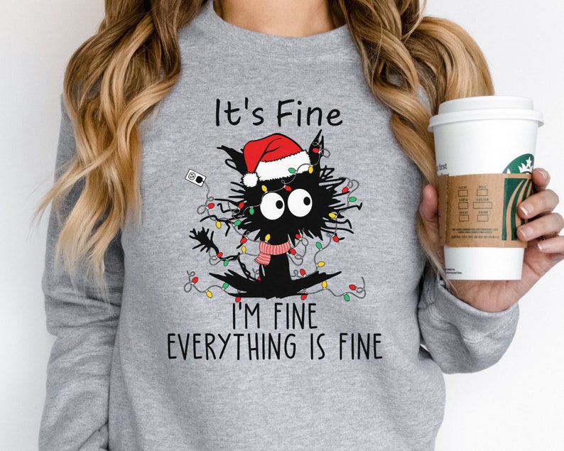 May include: A light gray sweatshirt featuring a black cat tangled in Christmas lights and wearing a Santa hat and scarf. The text "It's Fine, I'm Fine, Everything is Fine" is printed below the cat.