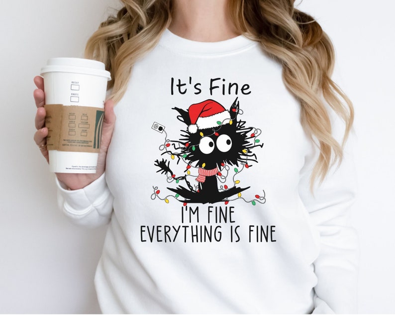 May include: White sweatshirt featuring a cartoon black cat tangled in Christmas lights and wearing a Santa hat and scarf. Text reads "It's Fine, I'm Fine, Everything is Fine."