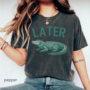 Op de afbeelding: Een donkergrijs T-shirt met een groene alligator-afbeelding en het woord "LATER" in het groen.