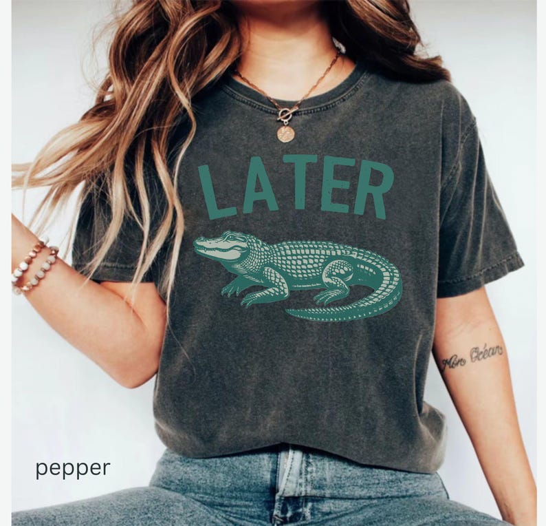 Op de afbeelding: Een donkergrijs T-shirt met een groene alligator-afbeelding en het woord "LATER" in het groen.