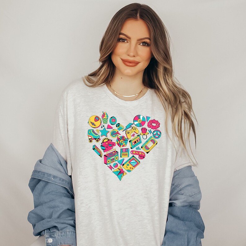 May include: A white t-shirt with a large heart-shaped design featuring a variety of 80s and 90s icons, including roller skates, cassette tapes, video game controllers, and ice cream cones. The icons are in shades of pink, blue, yellow, and green.