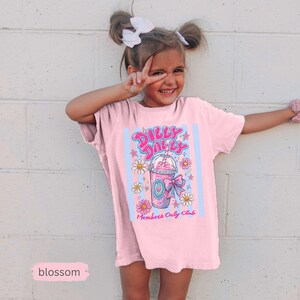 May include: A light pink t-shirt with a graphic that says "Dilly Dallys Members Only Club". The graphic features a pink drink with a straw, flowers, and stars. The shirt is worn by a young person.