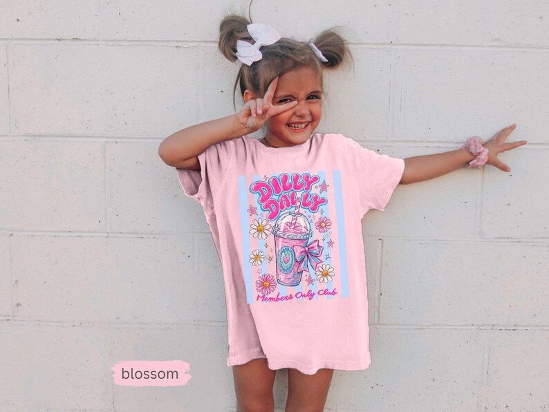 May include: A light pink t-shirt with a graphic that says "Dilly Dallys Members Only Club". The graphic features a pink drink with a straw, flowers, and stars. The shirt is worn by a young person.