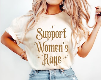 Op de afbeelding: Een wit T-shirt met de tekst "Support Women's Rage" gedrukt in bruine letters. De tekst is omringd door kleine sterren.