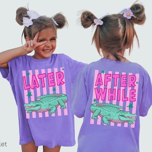 Puede incluir: Camisetas violetas con un gr&aacute;fico de un caim&aacute;n verde y el texto "LATER" en la parte delantera y "AFTER WHILE" en la trasera. El dise&ntilde;o incluye rayas rosas y estrellas verdes. Las camisetas las lleva un ni&ntilde;o.