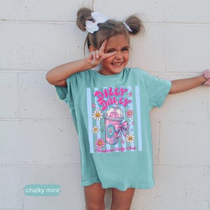 May include: A mint green t-shirt with a colorful graphic of a pink milkshake with daisies and the text "Dilly Dally" and "Members Only Club". The shirt is worn by a young person.