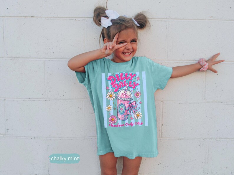 May include: A mint green t-shirt with a colorful graphic of a pink milkshake with daisies and the text "Dilly Dally" and "Members Only Club". The shirt is worn by a young person.