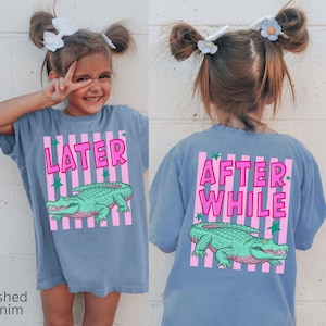 Puede incluir: Una camiseta azul con el texto "LATER" y un gr&aacute;fico de un caim&aacute;n verde en la parte delantera. La parte trasera de la camiseta tiene el texto "AFTER WHILE" y el mismo gr&aacute;fico de caim&aacute;n. La camiseta la lleva una ni&ntilde;a.