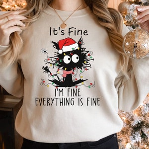 May include: Beige sweatshirt with a graphic of a black cat wearing a Santa hat and tangled in Christmas lights. Text reads "It's Fine, I'm Fine, Everything is Fine."