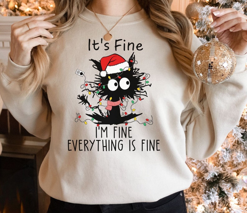 May include: Beige sweatshirt with a graphic of a black cat wearing a Santa hat and tangled in Christmas lights. Text reads "It's Fine, I'm Fine, Everything is Fine."