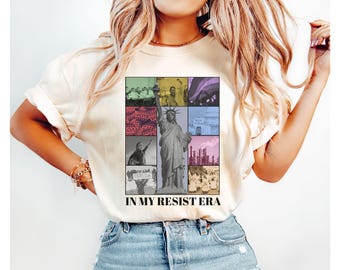 Puede incluir: Una camiseta blanca con un estampado gráfico de un collage de imágenes relacionadas con la Estatua de la Libertad, protestas y votaciones. El texto "IN MY RESIST ERA" está impreso debajo del collage.
