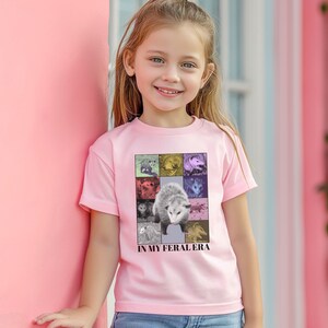 May include: A young person wearing a pink t-shirt with a graphic of an opossum and the text "In My Feral Era".
