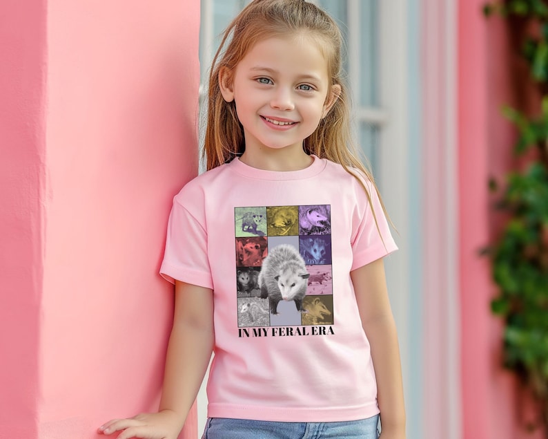 May include: A young person wearing a pink t-shirt with a graphic of an opossum and the text "In My Feral Era".