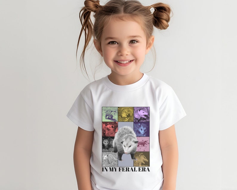May include: A white t-shirt with a colorful graphic of opossums. The graphic is a collage of different opossums in various poses. The text 'IN MY FERAL ERA' is printed below the graphic.