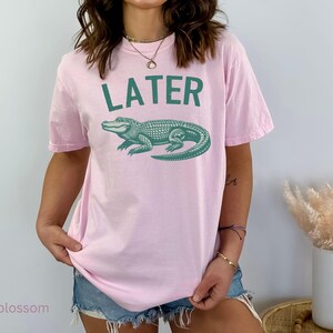Op de afbeelding: Roze T-shirt met een groene alligator-print en het woord "LATER" gedrukt op de voorkant.