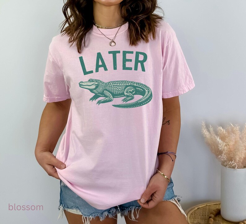 Op de afbeelding: Roze T-shirt met een groene alligator-print en het woord "LATER" gedrukt op de voorkant.