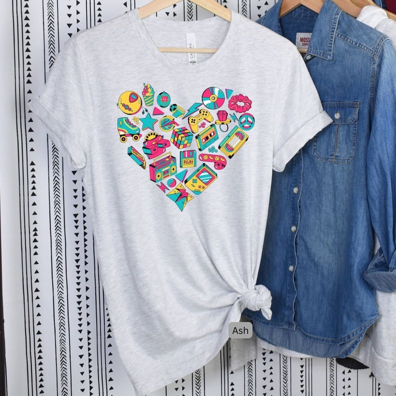 May include: A light gray t-shirt with a heart-shaped design featuring a variety of 80s and 90s icons, including a cassette tape, a boombox, a video game controller, a roller skate, and a Rubik's cube. The text "Ash" is visible on the bottom of the shirt.