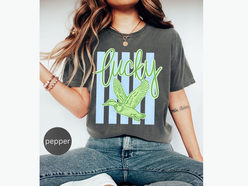 May include: A dark gray t-shirt with the word "Lucky" in green script above a green duck graphic. The word and duck are set against a light blue striped background. The shirt is a casual style.