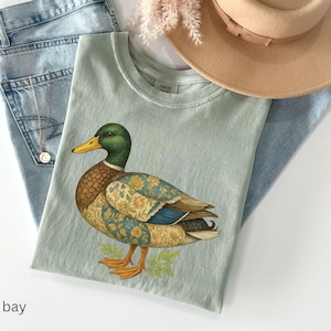Puede incluir: Camiseta verde claro con una ilustraci&oacute;n detallada de un pato. El pato tiene cabeza verde, cuerpo marr&oacute;n y alas con estampado floral. La camiseta est&aacute; sobre unos vaqueros azul claro y un sombrero beige.