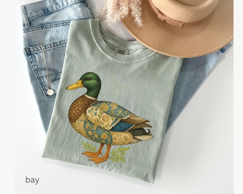 Puede incluir: Camiseta verde claro con una ilustraci&oacute;n detallada de un pato. El pato tiene cabeza verde, cuerpo marr&oacute;n y alas con estampado floral. La camiseta est&aacute; sobre unos vaqueros azul claro y un sombrero beige.