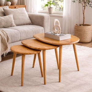 May include: A set of three wooden nesting tables with oval tops and tapered legs. The tables are arranged in a staggered configuration on a light-colored rug. A sofa and decorative items are in the background, creating a warm, inviting living space.