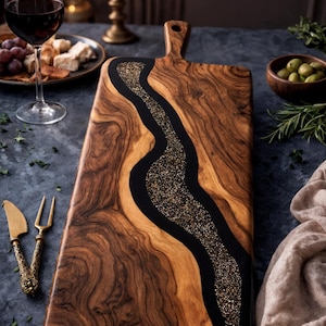 May include: A wooden serving board with a black resin river filled with gold glitter. The board has a handle. The image includes a glass of red wine, olives, and other food items.