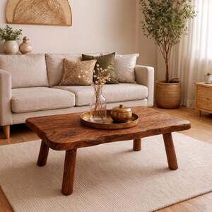 Könnte beinhalten: Ein rechteckiger Couchtisch aus massivem Holz mit natürlicher Kante und vier stabilen Beinen. Der Tisch steht auf einem hellbeigen Teppich vor einem beigen Sofa mit dekorativen Kissen. Ein goldenes Tablett mit einem goldenen Behälter und einer Glasvase mit getrockneten Blumen steht auf dem Tisch.