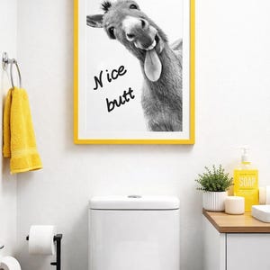 May include: A black and white framed print featuring a donkey with its tongue out, with the words "Nice butt" written on the image. The frame is yellow. The print is hanging above a white toilet in a bathroom.