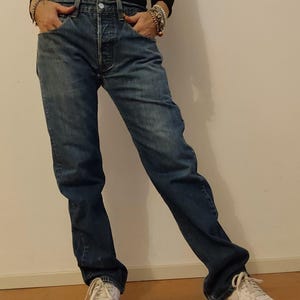 Può includere: Jeans di denim blu sbiadito con chiusura a bottoni e classico design a cinque tasche. I jeans sono indossati con scarpe da ginnastica bianche. L'aspetto generale è casual e confortevole.
