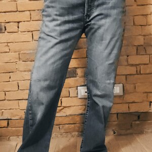 Könnte beinhalten: Verblasste schwarze Jeans mit Knopfleiste und klassischem Fünf-Taschen-Design. Die Jeans haben eine lockere Passform und werden mit den Händen in den Taschen getragen. Der Hintergrund ist eine Backsteinmauer.