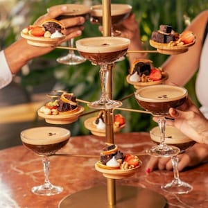 Peut inclure: Un présentoir doré à plusieurs niveaux contenant plusieurs martinis expresso dans des verres à pied. Chaque niveau contient également de petites assiettes en bois avec des desserts comme des brownies, des fraises et des noix. Les boissons ont une garniture crémeuse.