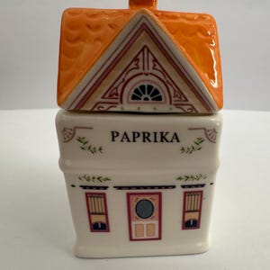 Op de afbeelding: Een huisvormige keramische paprikakruidenpot. Het heeft een oranje dak en een witte body met een deur en ramen. Het woord "PAPRIKA" staat op de voorkant.