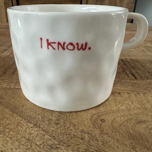 Può includere: Una tazza in ceramica bianca con una superficie testurizzata a fossette e un manico. La parte anteriore della tazza presenta il testo "I KNOW." stampato in un carattere semplice, rosso, in stile manoscritto. La tazza ha una forma cilindrica larga.