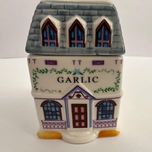 Op de afbeelding: Een keramische knoflookhouder in de vorm van een huis. Het huis heeft een grijs pannendak, gewelfde ramen en een deur. Het woord "GARLIC" staat op de voorkant gedrukt, met bloemmotieven. De houder is wit met blauwe, paarse en gele details.