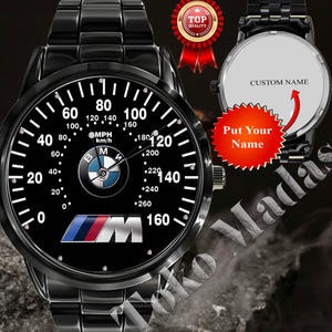 May include: Black wristwatch with a speedometer-style face featuring the BMW logo and the text "M". The watch face displays numbers up to 160 mph. The watch has a black metal band and the text "Put Your Name" is visible.
