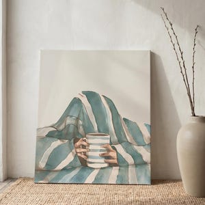 May include: A watercolor painting depicts a person holding a striped mug, covered by a blue and white striped blanket. The artwork is propped against a white wall, with a beige vase and a woven rug in the foreground.