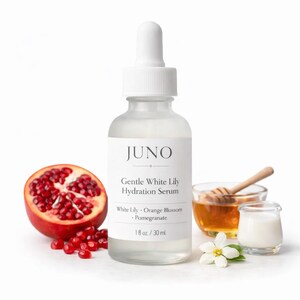 Puede incluir: Una botella de vidrio transparente de suero hidratante Gentle White Lily de Juno con un cuentagotas blanco. La botella est&aacute; junto a una granada cortada por la mitad, miel y un peque&ntilde;o plato de crema. La etiqueta enumera los ingredientes y el volumen de 30 ml.