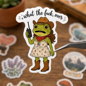 May include: A die-cut sticker featuring a green frog wearing a brown cowboy hat, a red bandana, and a white floral dress with brown cowboy boots. The frog holds a black walkie-talkie and the text above reads, what the fuck, over.